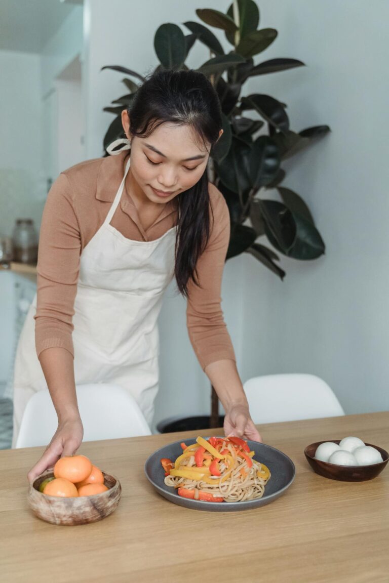Asian woman in a beige blouse serving a colorful noodle dish indoors at home.